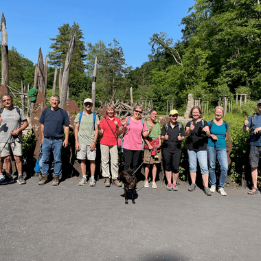 Gruppe von Wanderern mit Hunden posiert lächelnd vor einer Holzskulptur im Freien unter blauem Himmel.