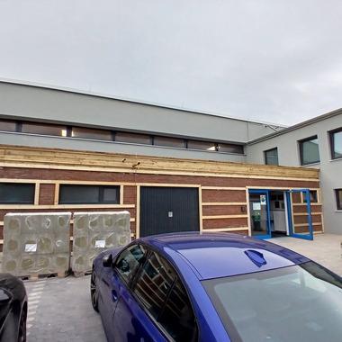 Außenansicht eines modernen Gebäudes mit Holzelementen, zwei Paletten und parkenden Autos im Vordergrund.