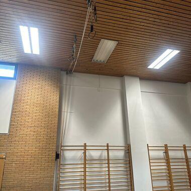 Seilzugmechanismus mit Ringen hängt an einer Holzbalkendecke in einem Sportraum mit Turnbänken.