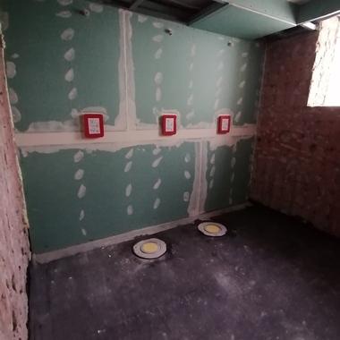 Rohbau eines Raumes mit grünen Wänden, zwei runden Bodenabläufen und roten Kabelkanälen an der Wand.