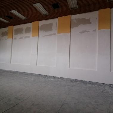 Unvollendete Wand mit gelben und weißen Abschnitten, daneben Baugerüst und abgedecktem Bodenbelag.