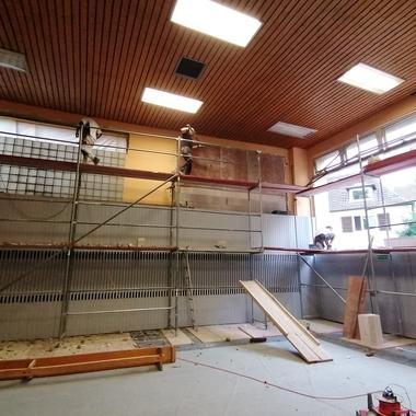 Blick in einen renovierungsbedürftigen Raum mit Baugerüst und arbeitenden Handwerkern an der Wand.