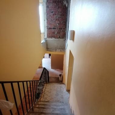 Blick auf eine Treppe mit schwarzem Geländer, gelben Wänden und freiliegendem Mauerwerk im oberen Bereich.