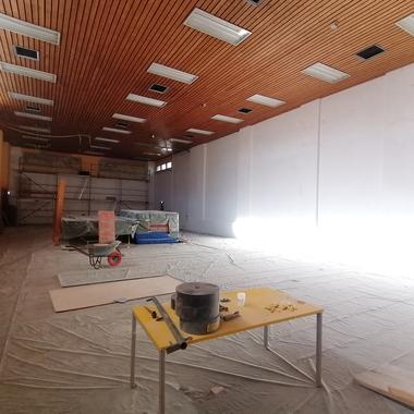 Innenansicht einer leerstehenden Turnhalle mit Holzdecke, Baustellenwagen und Materialien auf einem Abdeckplan.