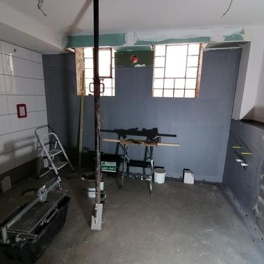 Baustelle mit einem geräumigen Raum, Leiter, Werkzeugen und grauen Wänden; Fensterblick auf Tageslicht.