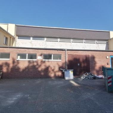 Außenansicht eines Gebäudes mit Backsteinfassade, blauen Fenstern und Baustellenmaterialien im Vordergrund.