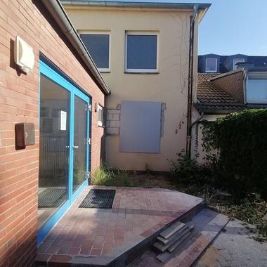 Eingangsbereich eines Gebäudes mit blauen Türen, einer baulichen Veränderung an der Wand und unangelegtem Plattenboden.