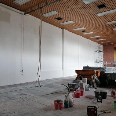 Innenansicht eines unvollendeten Raums mit einer weißen Wand, Baustellenmaterialien und einem Holzgerät auf einem Tisch.