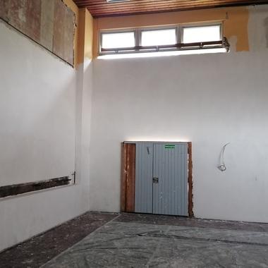 Leerer Raum mit weißen Wänden, einem grauen Tor und einem Fenster in der oberen Wand. Boden mit unebenem Belag.