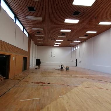Leere Sporthalle mit Holzfußboden, Wänden in Weiß und Holzverkleidung, Bauarbeiten im Gange.