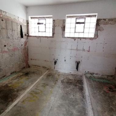 Leerer Raum mit unvollendeten Wänden, zwei Fenstern und sichtbaren Bodenmarkierungen für Sanitärinstallationen.