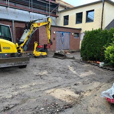 Baustelle mit gelbem Bagger, geräumtem Boden, Mauerwerk und Sträuchern in einem Wohnbereich.