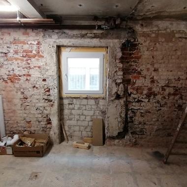 Blick in einen renovierungsbedürftigen Raum mit freiliegenden Ziegelwänden, einem Fenster und Baustellenmaterialien.