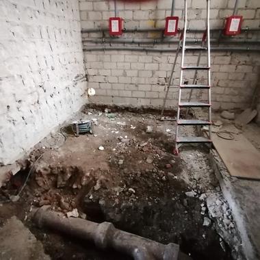 Blick in einen unvollendeten Raum mit freigelegtem Boden, einer Leiter und sichtbaren Rohren an der Wand.