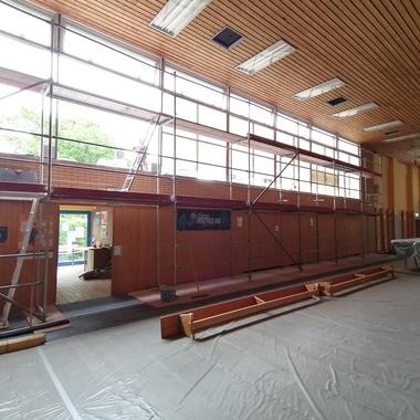 Blick auf eine Sporthalle mit Gerüst, Fenstern und abgedeckten Geräten auf einem grauen Bodenbelag.