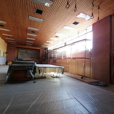 Innenansicht einer Sporthalle mit Holzdecke, Scaffolding und abgedeckten Geräten auf dem Boden. Tageslicht strömt hinein.