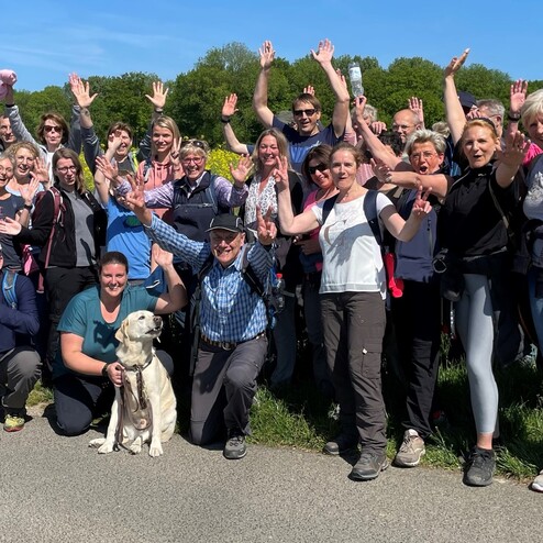 Gruppenselfie von Wanderern mit einem Hund, die fröhlich die Hände heben, vor gelben Rapsfeldern und blauem Himmel.