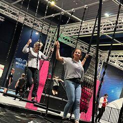 Zwei Frauen schwingen auf Seil-Schaukeln in einer Indoor-Trampolinanlage, während andere Besucher im Hintergrund aktiv sind.