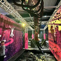 Zwei Kinder in einem Trampolinpark: eines schwingt an einer Röhre, das andere klettert an einer bunten Kletterwand.