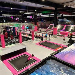 Trampolinpark mit mehreren Trampolinen, bunten Hindernissen und Kindern, die hüpfen und spielen. Beleuchtung in bunten Farben.