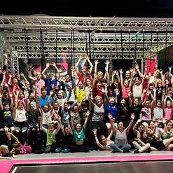 Gruppenspiel von Kindern und Betreuern in einem Trampolinpark, alle mit gehobenen Händen vor einem Trampolingelände.