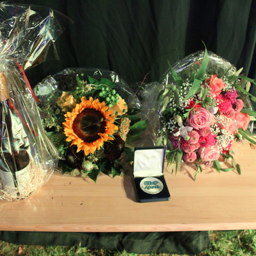 Geschenkset mit einer Flasche Wein in Folie, zwei Blumengestecken und einer Medaille auf einem Holztablett.