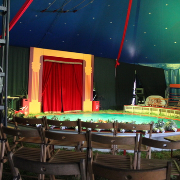 Zirkuszelt mit rotem Vorhang, grünem Boden und Holzstühlen um die Manege herum.