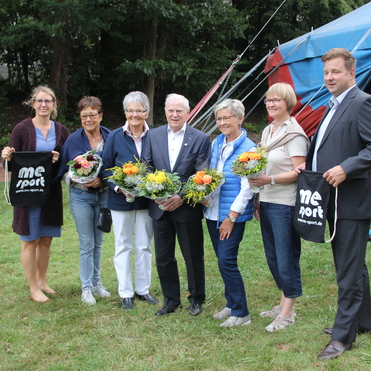 Gruppenfoto von sechs Personen mit Blumensträußen, die in einem Park vor einem Zelten stehen.