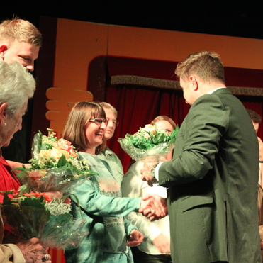 Gruppenfoto bei einer Veranstaltung, mehrere Personen schütteln Hände und halten Blumensträuße auf einer Bühne.