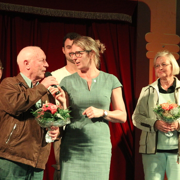 Eine Gruppe von sechs Personen steht auf einer Bühne, während ein älterer Mann mit Mikrofon spricht.