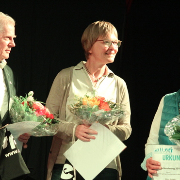 Drei Personen auf einer Bühne halten Urkunden und Blumensträuße, lächeln und schauen einander freundlich an.