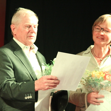 Zwei Personen halten Urkunden und Blumen auf einer Bühne; der Mann liest eine Seite in der Hand.