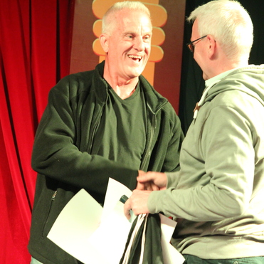 Zwei Männer schütteln sich lächelnd die Hände, einen Stapel Papiere zwischen sich haltend, vor rotem Vorhang.