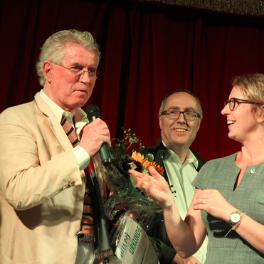 Zwei Personen stehen auf einer Bühne, während ein Mann mit Mikrofon einen Blumenstrauß hält und spricht.