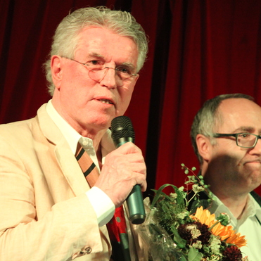 Mann mit Mikrofon hält einen Blumenstrauß, im Hintergrund ein anderer Mann, rote Vorhänge sichtbar.