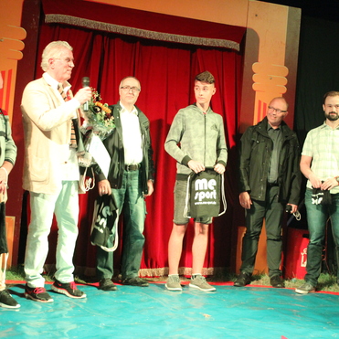 Gruppe von sechs Männern auf einer Bühne, einige halten Taschen, während ein Mann mit Mikrofon spricht. Rote Vorhänge im Hintergrund.