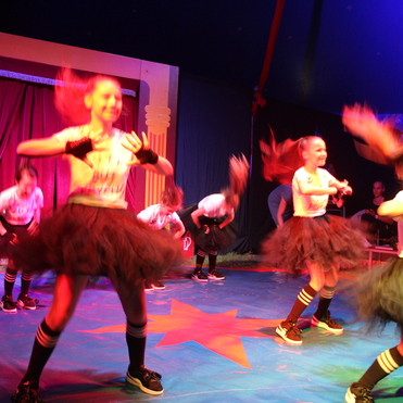 Tanzende Mädchen in Tüllröcken führen eine Choreografie unter bunten Lichtern in einem Zirkuszelt auf.