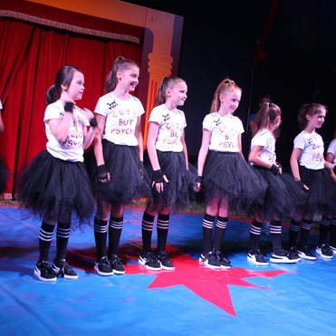 Gruppe von Mädchen in T-Shirts mit Aufschrift und schwarzen Tüllröcken steht auf einer bunten Bühne.