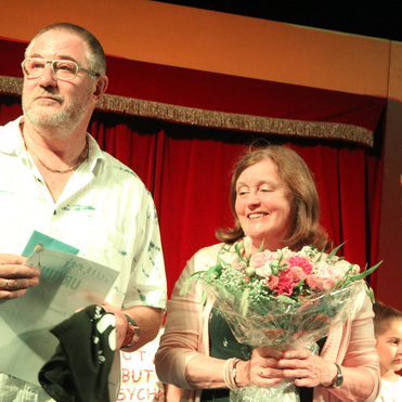 Zwei Personen stehen auf einer Bühne: ein Mann mit Dokumenten und eine Frau mit einem Blumenstrauß im Vordergrund.