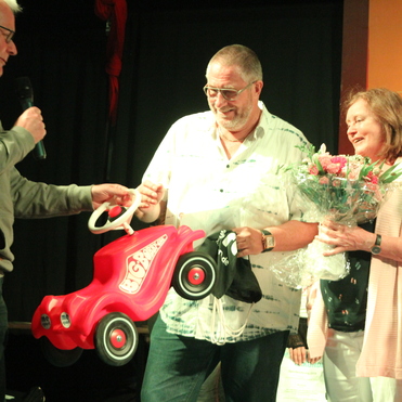 Mann überreicht einem anderen Mann ein rotes Spielzeugauto, während eine Frau mit Blumenstrauß lächelt.