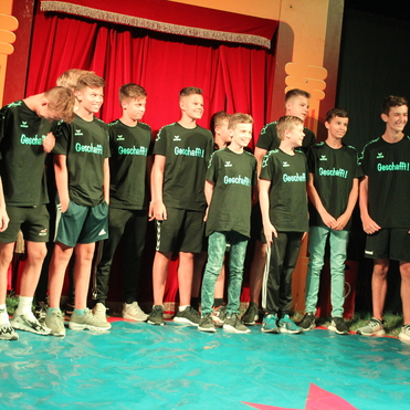 Gruppe von Jungen in schwarzen T-Shirts mit der Aufschrift "Geschafft!" steht lächelnd auf einer Bühne vor rotem Vorhang.