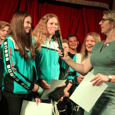 Junge Sportler:innen in blauen Jacken stehen auf einer Bühne, während eine Frau mit Mikrofon interviewt.