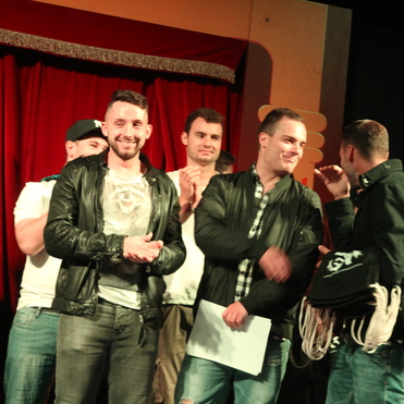 Fünf Männer stehen auf einer Bühne, einige applaudieren, ein roter Vorhang im Hintergrund.