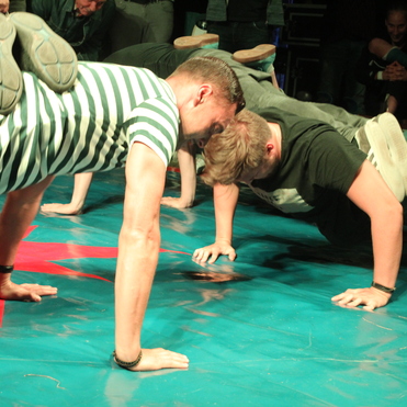 Zwei Personen machen Liegestütze auf einer blauen Matte, während andere im Hintergrund zuschauen.