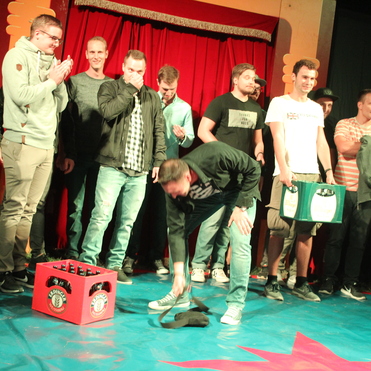 Eine Gruppe von Männern und Frauen steht auf einer Bühne vor einem roten Vorhang; ein Mann beugt sich zu einem schwarzen Gegenstand.