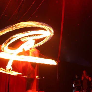 Zirkusaufführung mit Feuerspucker, der brennende Fackeln in spiralförmigen Mustern schwingt. Zuschauer im Hintergrund.