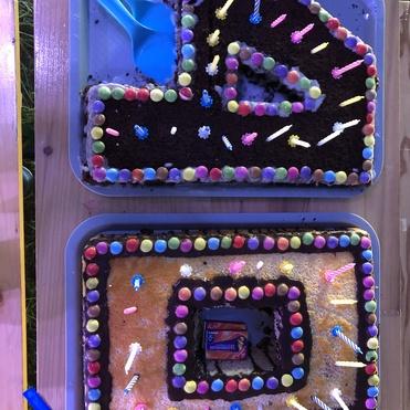 Bunte, rechteckige Kuchen in zwei Formen, dekoriert mit Bonbons und Kerzen, neben blauen Servierlöffeln auf einem Tisch.