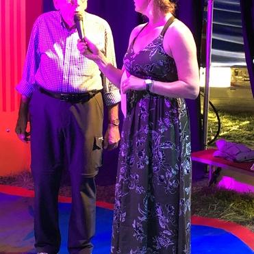 Ein älterer Mann spricht mit einer Frau, die ein Mikrofon hält, unter bunten Lichtern auf einer Bühne.