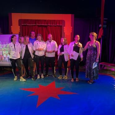 Sechs Frauen in Sportjacken stehen auf einem blauen Zirkusboden und sprechen mit einer Moderatorin in Abendkleid.