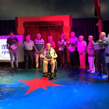 Mehrere Personen in einem Zirkuszelt sitzen oder stehen zusammen, eine Person im Rollstuhl hält Blumen. Grüne und rote Dekoration.